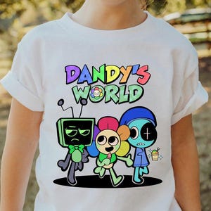 May include: White t-shirt featuring the text "Dandy's World" in colorful bubble letters above a cartoon illustration of three characters. The characters are a green TV-headed figure, a flower-like character, and a blue figure.