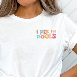 May include: White t-shirt with rolled-up sleeves featuring the text "I PEE IN POOLS" in colorful, playful lettering. The shirt is knotted at the waist, and the person is wearing black bottoms.