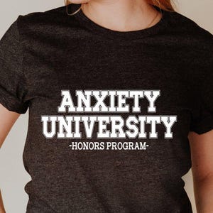 May include: Dark gray t-shirt with the text "ANXIETY UNIVERSITY -HONORS PROGRAM-" in white. The shirt is a crew neck style with rolled-up sleeves. The person in the image is wearing blue jeans and a black bracelet.