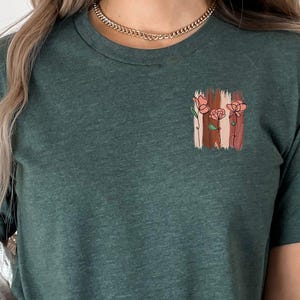 May include: Green t-shirt with a floral design on the chest. The design features pink flowers and vertical stripes in shades of brown and pink. The shirt is paired with blue jeans and a white cardigan.