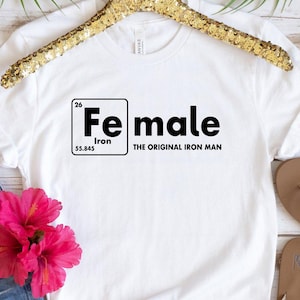 Puede incluir: Camiseta blanca con el texto "Fe male" en negro, con el s&iacute;mbolo qu&iacute;mico del hierro. La frase "The Original Iron Man" est&aacute; debajo. La camiseta est&aacute; combinada con pantalones cortos de mezclilla, gafas de sol, flores rosas y sandalias.