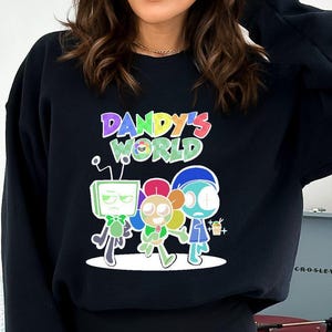 May include: Black sweatshirt featuring the text "Dandy's World" above a cartoon graphic of three characters. The graphic includes a TV-headed figure, a girl with pink hair, and a blue-skinned character. The sweatshirt is worn by a person.