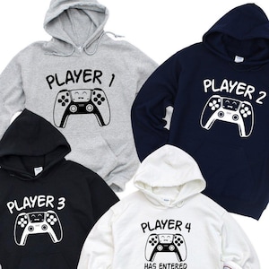 May include: Four hooded sweatshirts in gray, navy, black, and white. Each sweatshirt has a white graphic of a game controller and the words "PLAYER 1", "PLAYER 2", "PLAYER 3", and "PLAYER 4" printed on the front. White sneakers are at the bottom.