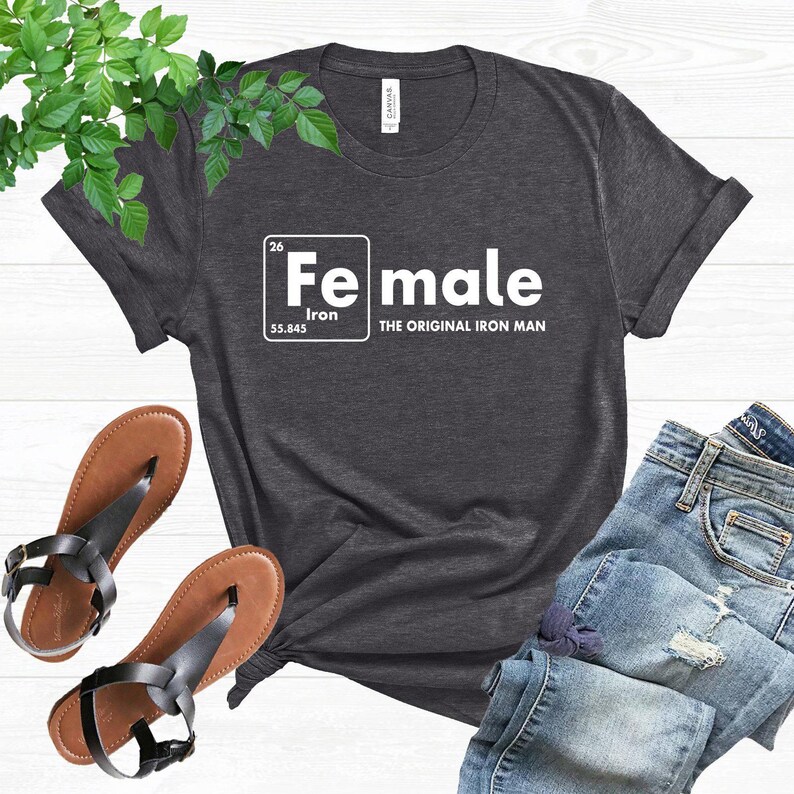 Puede incluir: Camiseta gris oscuro con la palabra "Female" en un estilo de tabla peri&oacute;dica, con "Iron" y "The Original Iron Man" debajo. La camiseta se combina con vaqueros azules y sandalias marrones.