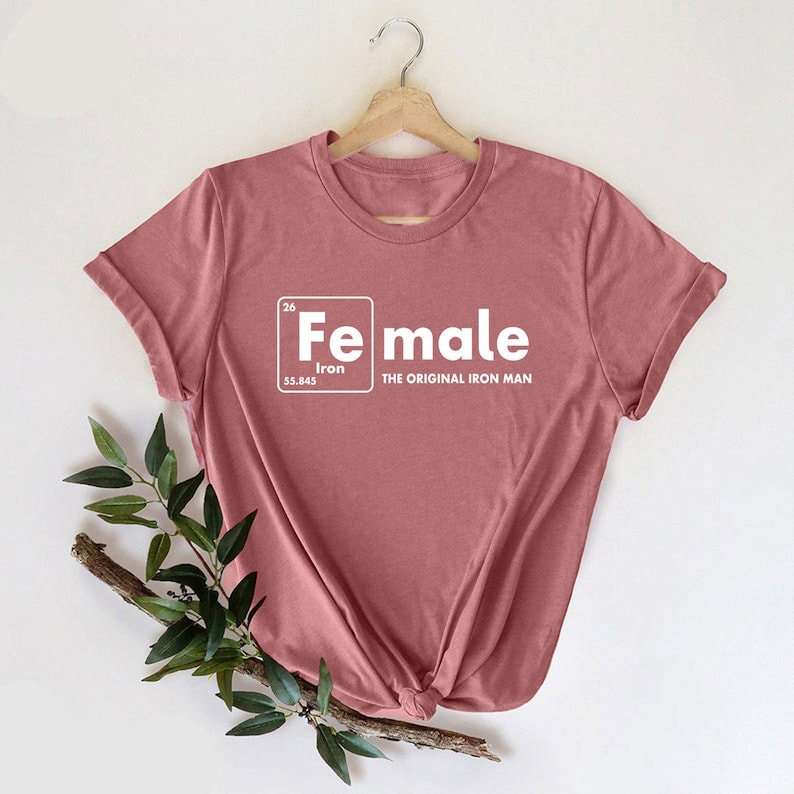 May include: A dusty rose t-shirt with rolled sleeves. The shirt features a white graphic that reads "Fe" with "Iron" below it, and "Female" below that, with the text "THE ORIGINAL IRON MAN" below the word "Female".