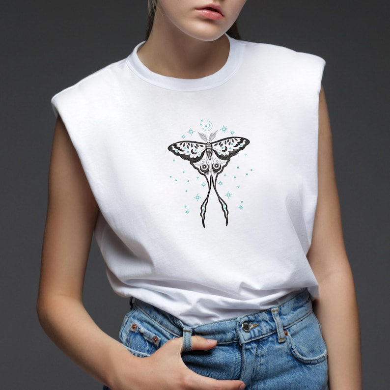 May include: White t-shirt featuring a black and white moth design with blue accents. The moth has long, thin wings and is surrounded by small blue stars.