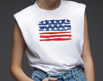 May include: White sleeveless t-shirt featuring a stylized American flag design. The flag graphic includes blue brushstrokes with white stars and red stripes. The shirt is paired with blue denim jeans.