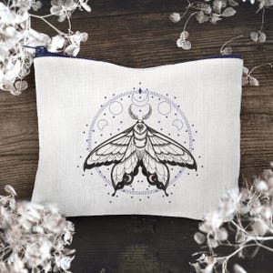 May include: A white linen zippered pouch with a black and white embroidered moth design. The moth is surrounded by a circle of moons and stars.