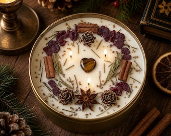 May include: A round, gold-colored metal tin candle with three lit wicks. The wax surface is decorated with cinnamon sticks, small pinecones, dried red berries, a star anise, and a smooth, heart-shaped brown gemstone in the center.