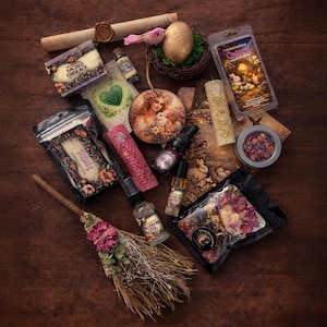 Op de afbeelding: Een assortiment spirituele en decoratieve items, waaronder een decoratieve bezem, kaarsen, etherische oliën, een decoratief ei in een nest en een verpakking gedroogde bloemen. De items zijn gerangschikt op een donker houten oppervlak.