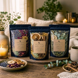 May include: Three black pouches of simmer pot blends with dried flowers, citrus, and spices. Each pouch has a label with the brand name and blend name: Soft Bloom, Golden Hearth, and Fresh Morning. A candle and decorative bowl are also visible.