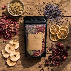 Black botanical simmer pot pouches arranged on a rustic wooden table with dried oranges, lavender, eucalyptus, cinnamon sticks, rose buds, herbs, and spices in a cozy home setting. Handcrafted home fragrance blends made with organic botanicals and natural ingredients.
