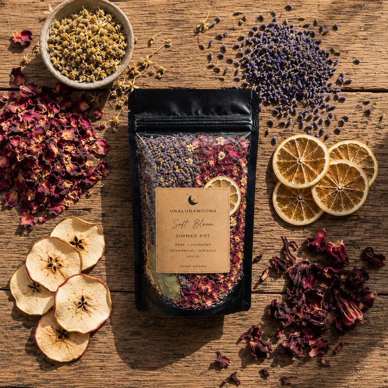 Black botanical simmer pot pouches arranged on a rustic wooden table with dried oranges, lavender, eucalyptus, cinnamon sticks, rose buds, herbs, and spices in a cozy home setting. Handcrafted home fragrance blends made with organic botanicals and natural ingredients.