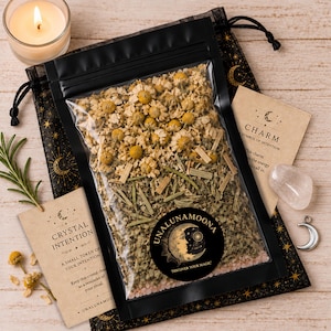 Op de afbeelding: Een doorzichtig plastic zakje gevuld met gedroogde kamillebloemen en groene kruiden, voorzien van een zwart rond etiket met de tekst UNALUNAMOONA DISCOVER YOUR MAGIC. Inclusief een zwart trekkoordzakje met gouden maan- en sterpatronen, twee beige kaarten met tekst, een helder kristal en een zilveren halve maan bedel.
