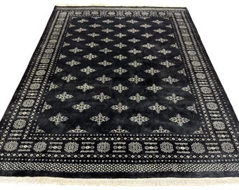 Puede incluir: Una alfombra rectangular negra con un patrón repetitivo de diamantes y flores en blanco roto. La alfombra tiene un borde detallado con diseños geométricos y flecos en los bordes. Adecuada para la decoración del hogar.