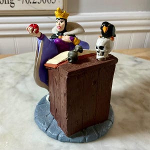 May include: A figurine of the Evil Queen from Snow White, wearing a crown and holding a red apple. The figurine is on a wooden desk with a skull and a raven. The base is a stone-like texture.