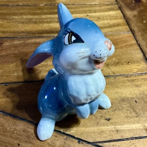 May include: A ceramic bunny figurine in shades of blue, with a pink nose and inner ears. The bunny has a smiling expression and is posed in a sitting position. The figurine is on a wooden surface.