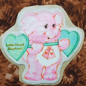 May include: A pink plush elephant Care Bear with a green heart that says "Lotsa Heart Elephant" and a blue heart. The Care Bear is holding a pink rattle with hearts on it.