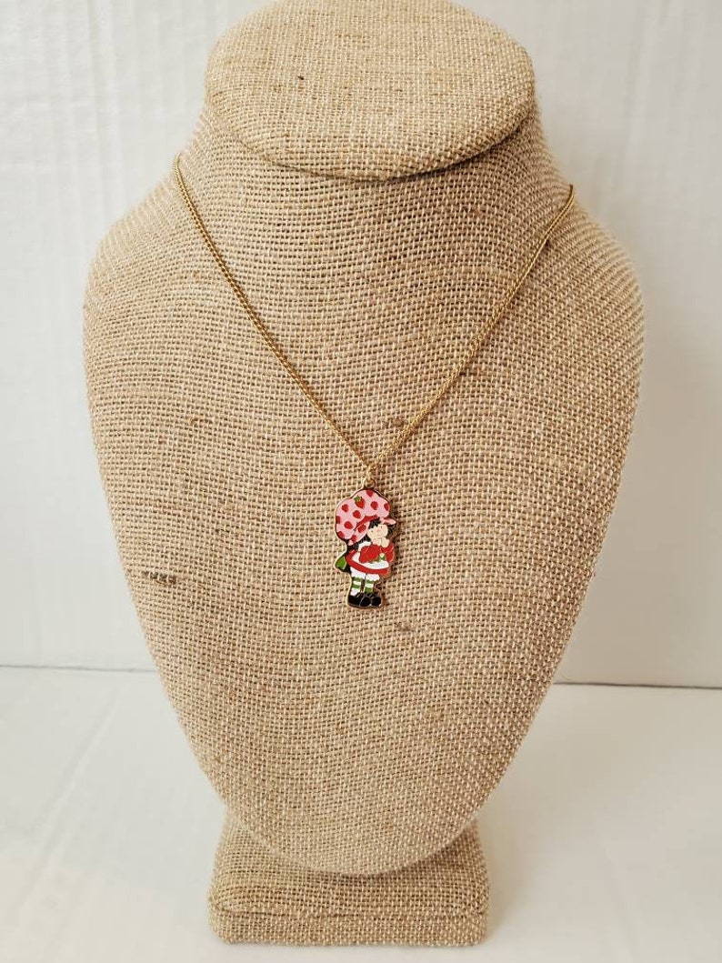 May include: A gold chain necklace with a red and white enamel charm of a girl holding a pink and white polka dot balloon.