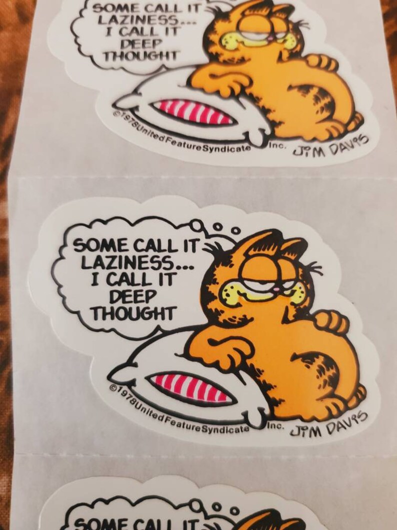 May include: A cartoon orange cat with a thought bubble above its head. The thought bubble says "SOME CALL IT LAZINESS... I CALL IT DEEP THOUGHT". The cat is lying on a striped pillow. The image is a sticker with the copyright information "&copy;1978 United Feature Syndicate Inc. JM DAVIS".