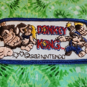 May include: A white embroidered patch with a blue border featuring the characters from the Donkey Kong video game. The text "Donkey Kong" is in red and blue lettering with a yellow star. The text "©1982 Nintendo" is in black lettering.