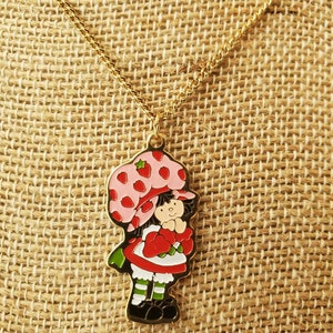May include: A gold chain necklace with a charm of Strawberry Shortcake. The charm is a cartoon image of Strawberry Shortcake with a pink hat and red dress.