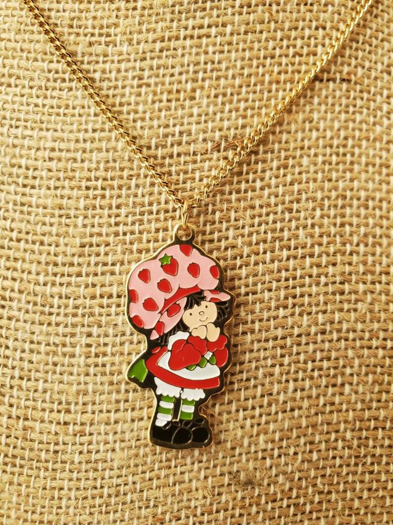 May include: A gold chain necklace with a charm of Strawberry Shortcake. The charm is a cartoon image of Strawberry Shortcake with a pink hat and red dress.