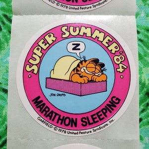 Op de afbeelding: Een vel met ronde stickers met Garfield de kat die slaapt. De stickers hebben een roze rand met de woorden "SUPER SUMMER '84" en "MARATHON SLEEPING". Garfield ligt onder een deken met een "Z" in een tekstballon.