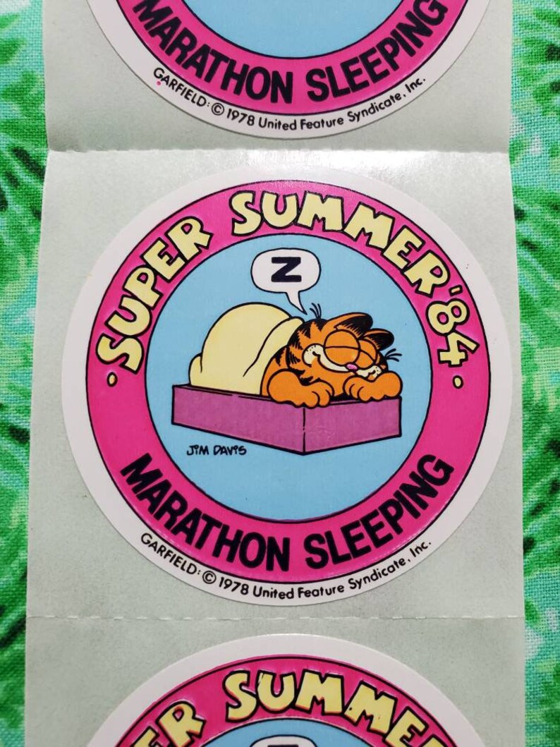 Op de afbeelding: Een vel met ronde stickers met Garfield de kat die slaapt. De stickers hebben een roze rand met de woorden "SUPER SUMMER '84" en "MARATHON SLEEPING". Garfield ligt onder een deken met een "Z" in een tekstballon.