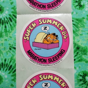 Op de afbeelding: Een vel met ronde stickers met Garfield de kat die slaapt. De stickers hebben een roze rand met de woorden "SUPER SUMMER '84" en "MARATHON SLEEPING". Garfield is oranje en geel, slapend onder een deken.