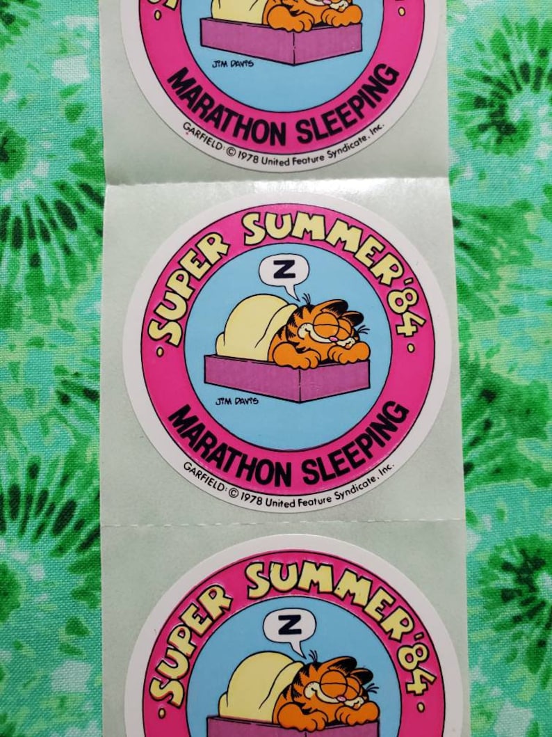 Op de afbeelding: Een vel met ronde stickers met Garfield de kat die slaapt. De stickers hebben een roze rand met de woorden "SUPER SUMMER '84" en "MARATHON SLEEPING". Garfield is oranje en geel, slapend onder een deken.
