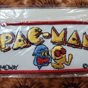 May include: A white embroidered patch with the words "PAC-MAN" in yellow and red lettering. The patch features a yellow Pac-Man character with a red mouth and a blue ghost character. The patch also includes the words "MIDWAY" and "1981" in black lettering.