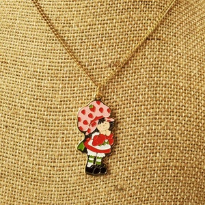 May include: A gold chain necklace with a charm of Strawberry Shortcake, a cartoon character with a red dress, green shoes, and a pink hat with strawberries on it.