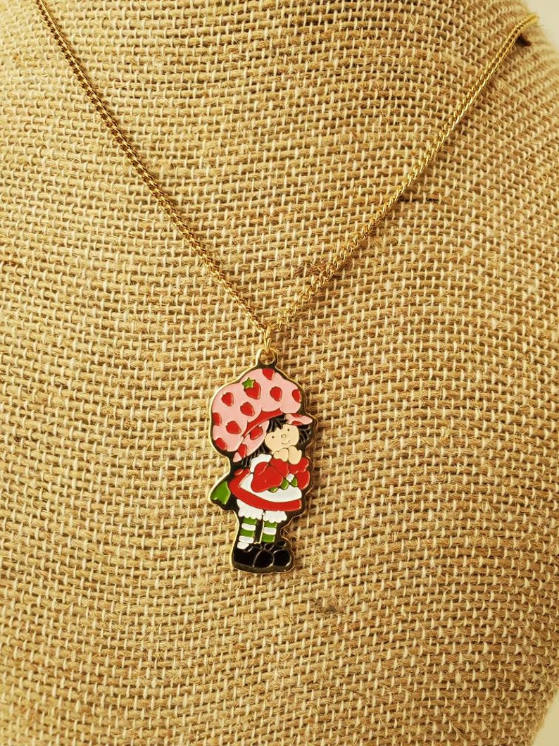 May include: A gold chain necklace with a charm of Strawberry Shortcake, a cartoon character with a red dress, green shoes, and a pink hat with strawberries on it.
