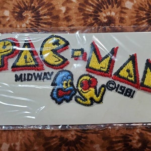 May include: A vintage Pac-Man iron-on patch set. The set includes the Pac-Man logo, two ghosts, and the Midway logo. The patch set is yellow, red, and black with the text "MIDWAY" and "©1981".