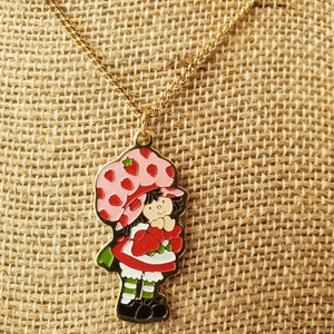 May include: A gold chain necklace with a charm of Strawberry Shortcake. The charm is a cartoon image of Strawberry Shortcake with a pink hat and red dress.