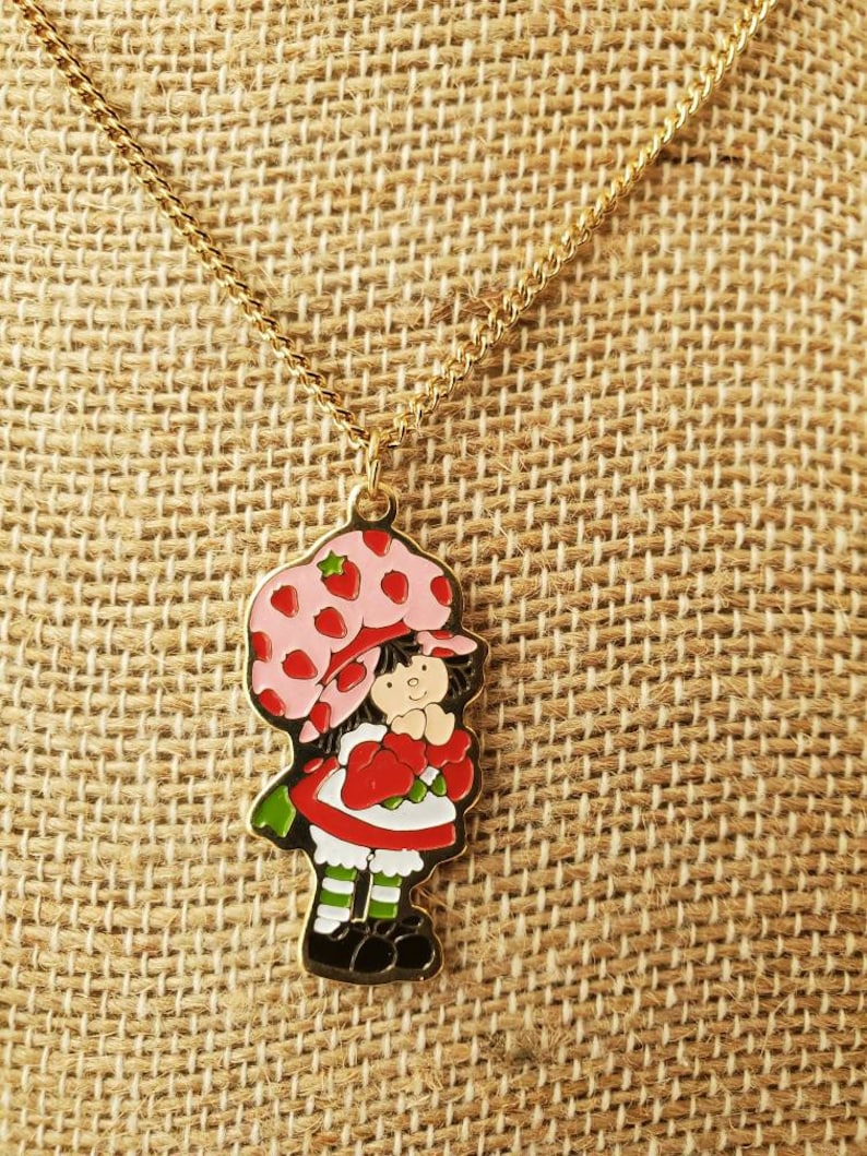 May include: A gold chain necklace with a charm of Strawberry Shortcake. The charm is a cartoon image of Strawberry Shortcake with a pink hat and red dress.