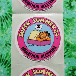 Op de afbeelding: Een vel met ronde Garfield-stickers. Elke sticker toont Garfield die onder een deken slaapt met een gedachtewolk met de letter "Z". De stickers hebben een roze rand met de woorden "Super Summer '84" en "Marathon Sleeping".