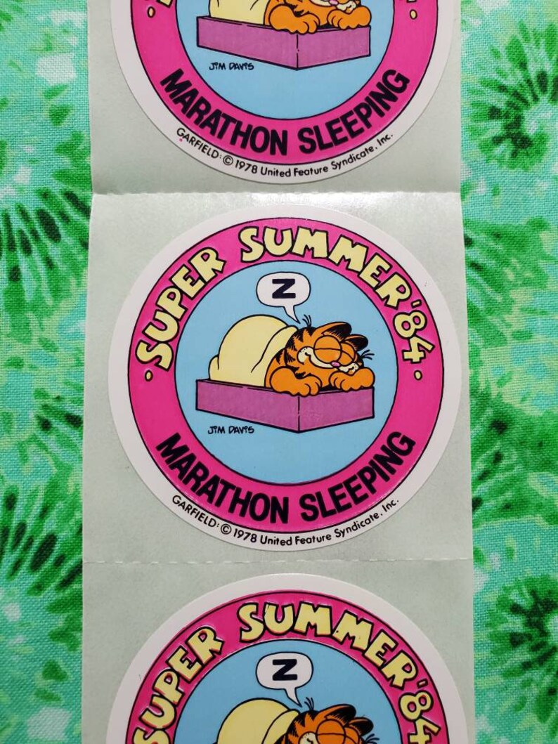 Op de afbeelding: Een vel met ronde Garfield-stickers. Elke sticker toont Garfield die onder een deken slaapt met een gedachtewolk met de letter "Z". De stickers hebben een roze rand met de woorden "Super Summer '84" en "Marathon Sleeping".