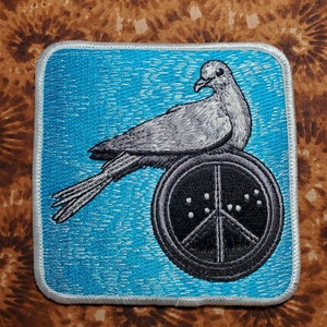 May include: A blue embroidered patch with a white dove perched on a black peace sign. The dove has a gray body and a black eye.