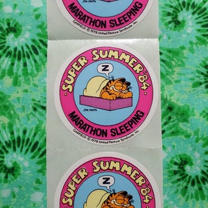 Op de afbeelding: Garfield-stickers met de stripkat die in bed slaapt. De stickers hebben een roze en blauw cirkelvormig ontwerp met de tekst "Super Summer '84" en "Marathon Sleeping". De stickers zitten op een vel.