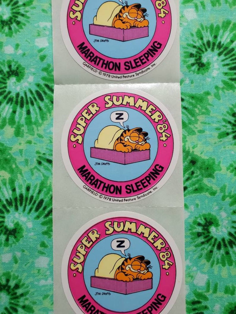 Op de afbeelding: Garfield-stickers met de stripkat die in bed slaapt. De stickers hebben een roze en blauw cirkelvormig ontwerp met de tekst "Super Summer '84" en "Marathon Sleeping". De stickers zitten op een vel.