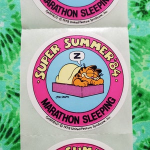 Op de afbeelding: Een vel met ronde stickers met Garfield de kat die slaapt. De stickers hebben een roze rand met de woorden "SUPER SUMMER '84" en "MARATHON SLEEPING". De cartoonkat is oranje en geel en slaapt op een paars bed met een gele deken.