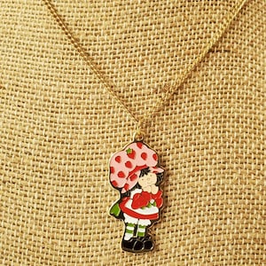 May include: A gold chain necklace with a charm of Strawberry Shortcake. The charm is red, white, and green with a pink and white hat.