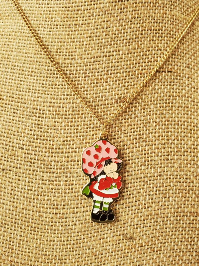 May include: A gold chain necklace with a charm of Strawberry Shortcake. The charm is red, white, and green with a pink and white hat.