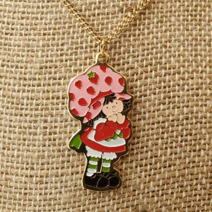 May include: A gold chain necklace with a charm of Strawberry Shortcake. The charm is a cartoon image of Strawberry Shortcake wearing a pink and red hat and dress. She is holding a red strawberry.