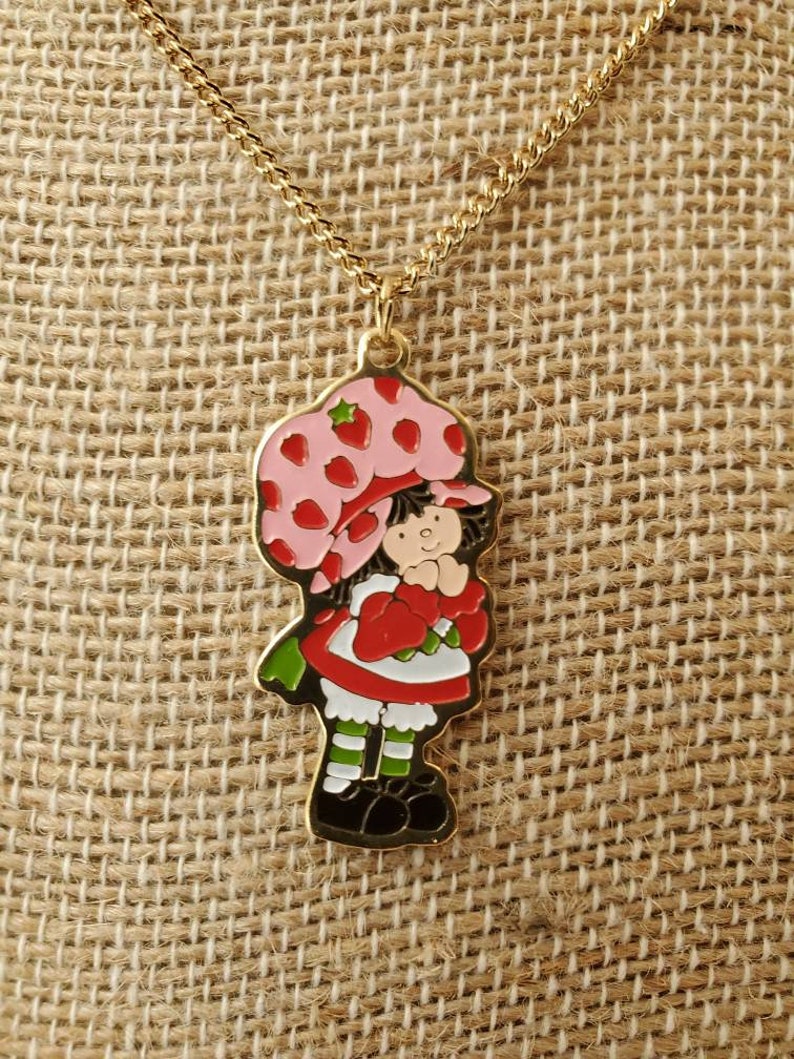 May include: A gold chain necklace with a charm of Strawberry Shortcake. The charm is a cartoon image of Strawberry Shortcake wearing a pink and red hat and dress. She is holding a red strawberry.