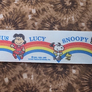 May include: Light blue ribbon featuring characters Linus, Lucy, and Snoopy with a rainbow design. The ribbon has the text "LINUS", "LUCY", and "SNOOPY". The ribbon is against a brown and white patterned background.