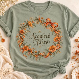 May include: A sage green t-shirt featuring a circular wreath graphic composed of orange flowers, green leaves, a coiled snake, and an orange ribbon. The text "Acquired Taste" is printed in a dark brown script font in the center of the wreath.