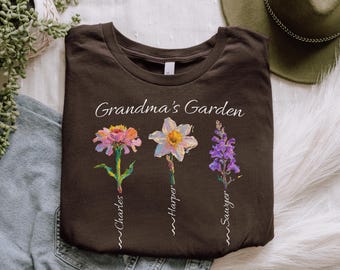 Può includere: Una maglietta marrone con la scritta "Grandma's Garden" e tre fiori ad acquerello. Ogni fiore ha un nome: Charles, Harper e Sawyer. I fiori sono una zinnia rosa, un narciso bianco e una bocca di leone viola.